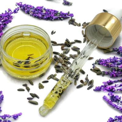 small bowl of lavender jojoba oil and lavender flowers sitting next to a dropper filled with lavender jojoba oil. All lying on a bed of lavender buds