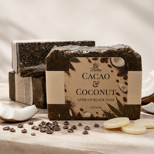 Three bars of Fuller Cacao & Coconut African Black Soap arranged on a light stone surface with coconut halves, scattered cacao beans, and sliced coconut. The front bar has a kraft paper label with product details, and the other bars are wrapped in dark paper.